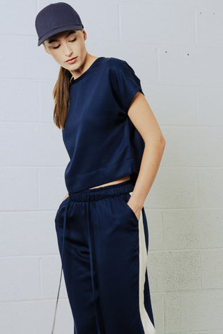 Woman in front of white brick wall wearing navy blue baseball cap, short sleeve t-shirt and draw-string pants with white tuxedo stripe. 