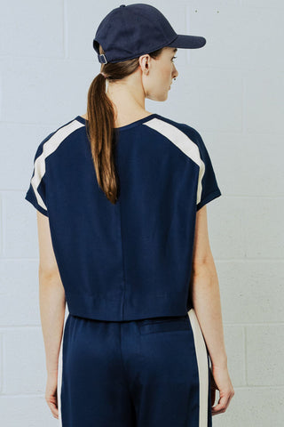 Woman in front of white brick wall wearing navy blue baseball cap, short sleeve t-shirt and pants with white tuxedo stripe. 