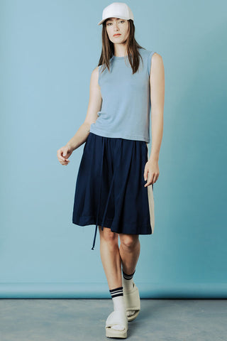 Woman in front of blue background wearing white cap, blue tank top and navy blue knee length shorts with ivory stripe down side. 
