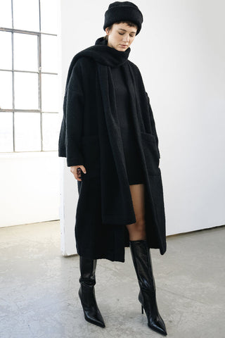 A model wearing a long black coat, black beret, black dress, and black pointed-toe heeled boots, standing in a bright studio with a window and concrete floor.
