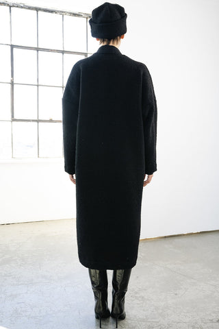 A person viewed from behind wearing a long black coat, black beanie, and black boots in a bright studio with a large window and white wall.
