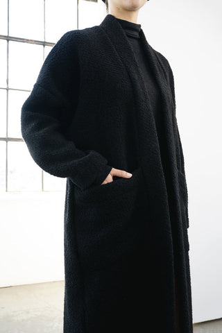 A person wearing a long black textured coat over a black turtleneck, standing in a bright indoor setting with a window in the background.