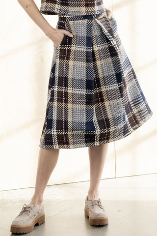 A person wearing a pleated A-line skirt in a navy, beige, and brown plaid pattern, paired with beige lace-up shoes, standing in a brightly lit indoor space.