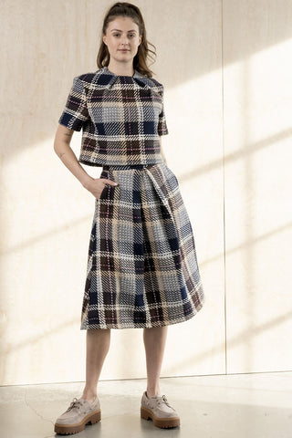 A woman models a matching short-sleeve cropped top and A-line midi skirt set in a navy, beige, and brown plaid, styled with beige shoes against a warm beige wall with window shadows.