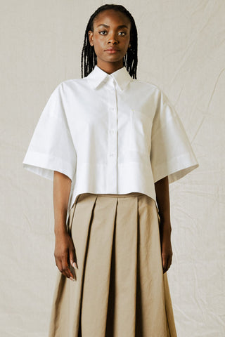 Woman in front of beige background wearing white button-up shirt with wide sleeves and tan pleated skirt. 