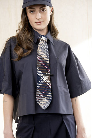 Person wearing a navy cropped short sleeve button up shirt with a patterned tie against a neutral background