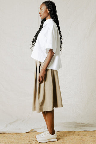 Woman in front of beige background wearing white button-up shirt with wide sleeves and tan pleated skirt. 