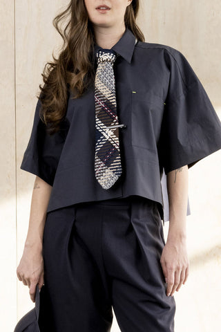A model wearing a dark navy blue short-sleeve button-up shirt with a matching dark navy pleated trouser, styled with a patterned plaid tie.