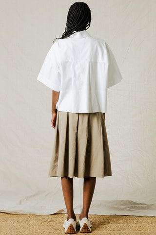 Back view of woman in front of beige background wearing white button-up shirt with wide sleeves and tan pleated skirt. 