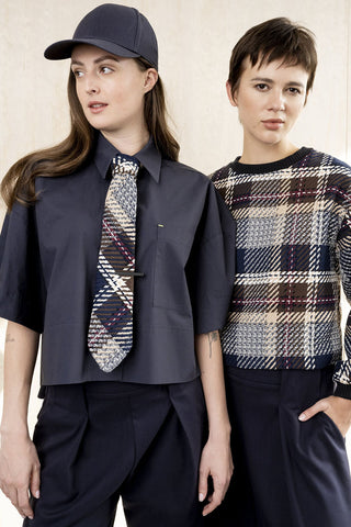 Two models wearing a dark navy blue fashion ensemble, featuring a cropped button-up shirt with a patterned tie, matching navy trousers, and a navy baseball cap, alongside a plaid long-sleeve top.