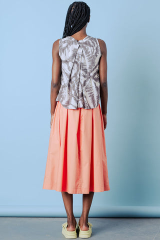 Back view of woman wearing patterned shirt and pleated coral skirt in front of blue background. 