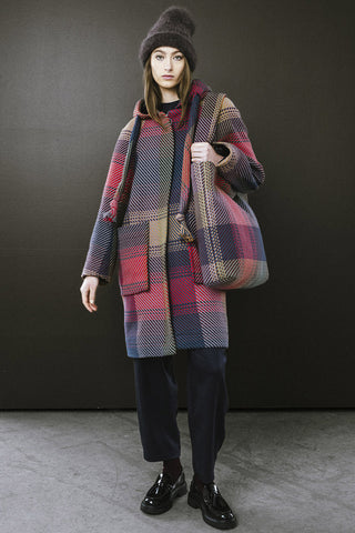 Woman standing in front of dark wall, wearing navy and red plaid zip up Morrison Coat by BodyBag and matching bag. 