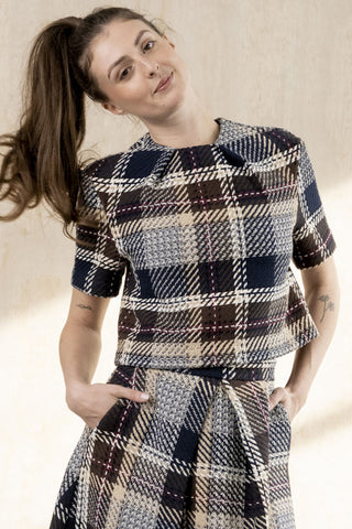A woman wearing a short-sleeve plaid top with pleats at neck, and matching skirt, photographed against a warm beige wall.