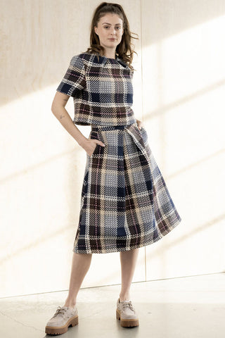A model wears  a matching short-sleeve blouse and pleated A-line skirt set in a navy, beige, and brown plaid tweed fabric, paired with beige lace-up shoes, set against a warm beige wall. 