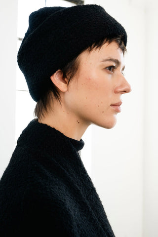 A side-profile portrait of a woman with short dark hair wearing a black textured toque and matching black top against a bright white background.