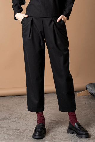 A woman wearing loose black cropped trousers with pleated front details, paired with a black top, burgundy socks, and black leather loafers, standing against a warm beige studio backdrop.
