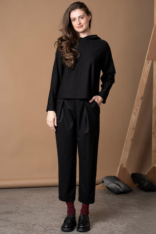 A woman wearing a black long-sleeve cropped hoodie and black pleated cropped trousers, styled with dark red socks and black loafers, against a warm brown studio backdrop.