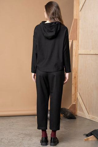 Awoman viewed from the back wearing a black hooded sweatshirt and black cropped pants, standing in a studio with a beige backdrop and wooden paneling.