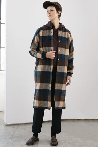 A model wearing a long-line plaid coat styled with black trousers, brown loafers, and a brown cap in a minimalist studio setting.
