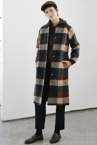 A model wearing a long-line plaid coat styled with black trousers, brown loafers, and a brown cap in a minimalist studio setting.