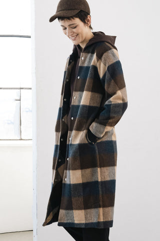 A model wearing a long-line plaid coat styled with brown hoodie, black trousers, and a brown cap in a minimalist studio setting.