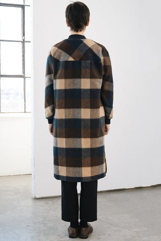 A person viewed from behind wearing a long plaid coat in black, brown, and beige, paired with black trousers and brown shoes, standing in a bright white room with a window.