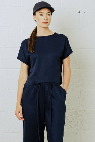 Woman in front of white brick wall wearing navy blue baseball cap, short sleeve t-shirt and draw-string pants. 