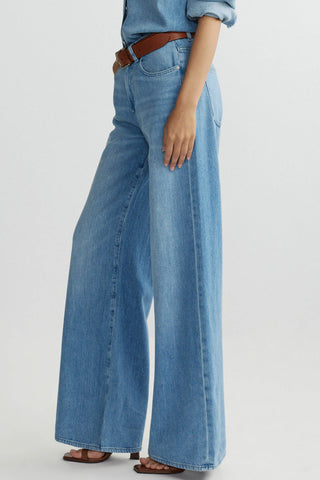Side view of woman in front of plain background wearing mid-blue wash baggy wide leg jeans with brown leather belt. 