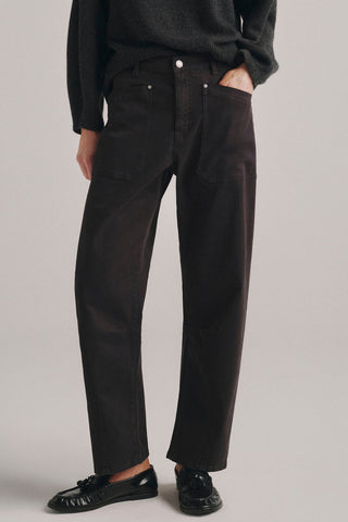 A person wearing dark brown high rise barrel fit jeans, styled with a dark gray knit sweater and black leather loafers, photographed against a neutral light gray studio background.