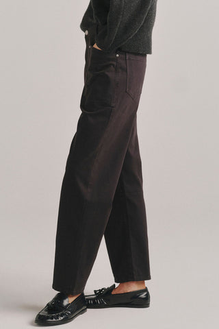 A person shown from the waist down wearing dark brown barrel leg jeans, a dark knit sweater, and black leather loafers against a plain light gray studio background.