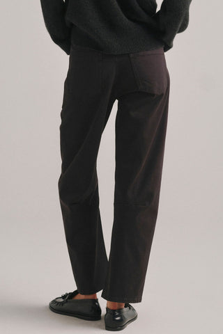A person shown from the back, waist down wearing dark brown barrel fit  jeans, a dark knit sweater, and black loafers against a plain light gray studio background.