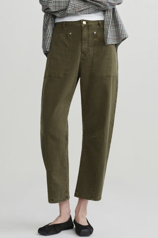 A person shown from waist down wearing olive green cropped barrel leg jeans with black flats, shown against a light gray studio background.