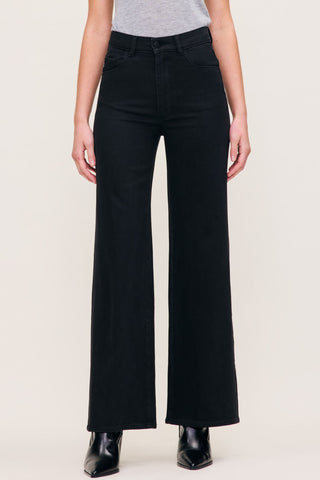 A model wearing black wide-leg jeans with a gray top and black pointed-toe boots, shown from the waist down against a light beige studio background.