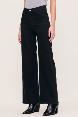 A model wearing black wide-leg jeans with a gray top and black pointed-toe ankle boots, shown from the waist down against a light beige studio background.