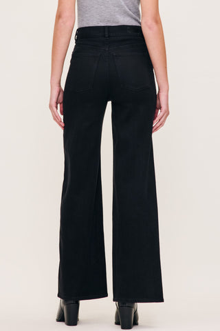 Rear view of a model wearing black wide-leg jeans with a gray top and black heeled boots, set against a plain off-white studio background.