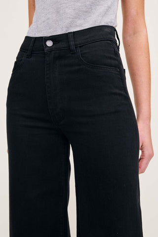 Close-up of a person wearing black high-waisted jeans with a gray top, showing the front waistband, button, and front pockets against a plain light background.