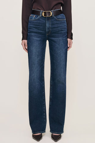 A model is shown from the waist down wearing dark blue straight-leg jeans with a gold-tone belt buckle, a dark brown long-sleeve top, and dark brown pointed-toe heels against a light beige studio background.