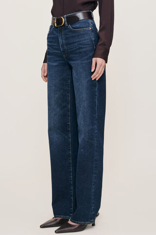 A model wearing dark blue wide-leg jeans with a black belt, paired with a dark brown long-sleeve top and black pointed-toe heels, shown from the waist down against a light beige studio background.