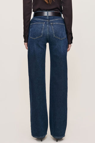 Rear view of a model wearing dark blue wide-leg denim jeans with a black leather belt, paired with a dark brown long-sleeve top and black heels, against a light beige studio background.