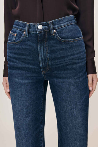 A close-up of a model wearing dark blue denim jeans with a high waist and straight-leg fit, paired with a dark brown long-sleeve top against a light beige background.