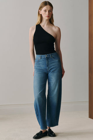 Woman in front of plain background wearing black one shoulder top tucked into blue wash barrel cut jeans. 