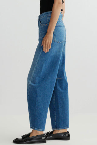Side view of woman in front of plain background wearing blue wash barrel leg jeans and black loafers. 