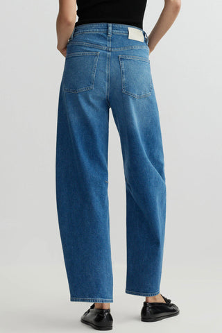 Back view of woman in front of plain background wearing blue wash barrel leg jeans and black loafers. 