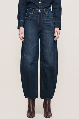 A model shown waist down wearing dark blue barrel leg jeans with brown ankle boots, against a light beige studio background.