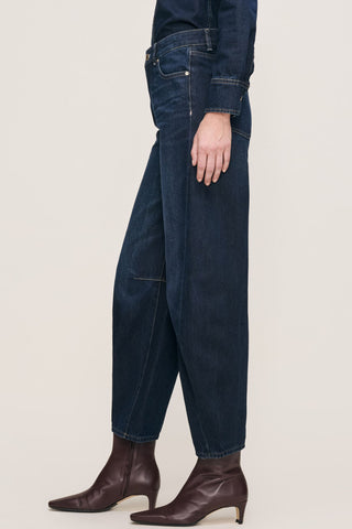 A waist down side view of a person wearing dark blue barrel leg with dark brown pointed-toe ankle boots against a plain light beige studio background.