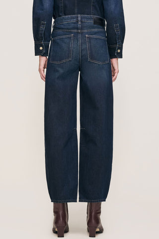 Rear view of a model wearing dark blue barrel leg jeans with brown leather ankle boots against a light beige studio background.