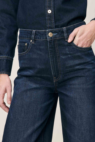 Close-up of a person wearing dark indigo blue denim jeans with a buttoned shirt, showing the front waistband, belt loops, front pockets, and hand in pocket.