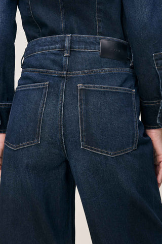 Close-up back view of a person wearing dark blue denim jeans with a black leather brand patch on the waistband, shown against a light neutral background.