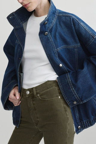 A close-up of a person wearing an oversized dark blue denim jacket over a white top and olive green pants, photographed against a light gray studio background.