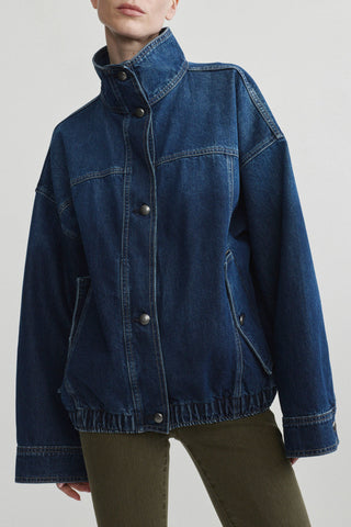 A model wearing a dark blue denim jacket with a high collar and button front, paired with olive green pants, against a light gray studio background.